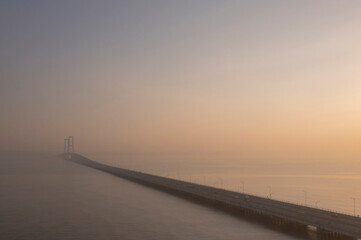 Fototapeta premium Aerial view of the long Suramadu Bridge stretching across the ocean during a hazy sunrise, with the bridge structure fading into the golden misty horizon