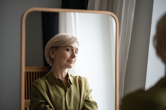 Beautiful smiling middle aged woman standing and looking in mirror with positive expression, blonde lady enjoying own reflection and outlook, Female looks satisfied with new haircut and clothing.