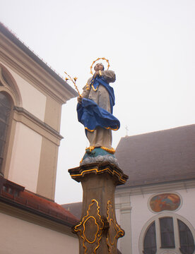 Religious statue of a saint with halo and blue robe on a decorated pedestal in a historic church courtyard, surrounded by traditional Bavarian architecture in foggy weather, 30 November 2025 Dillingen