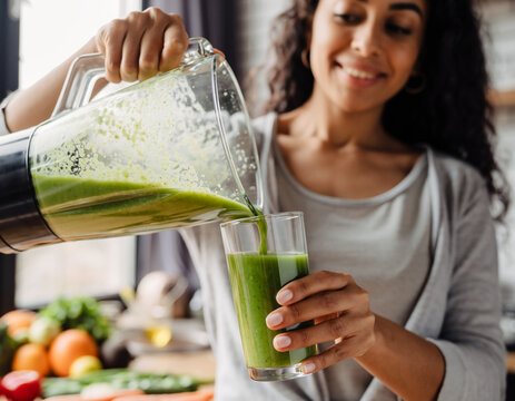 Young woman pouring a vibrant green smoothie into a clear glass, healthy lifestyle concept, fresh ingredients in background - Powered by Adobe