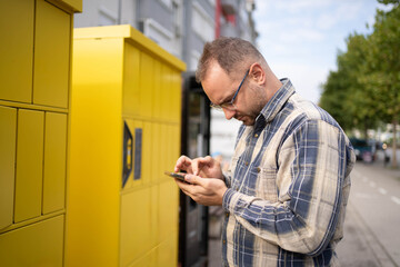Man interacting with smartphone near yellow delivery lockers in urban setting during daylight hours