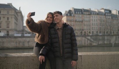A romantic couple takes a selfie photo with a mobile phone on a bridge over the Seine River in Paris France with city buildings in the background on a sunny day. This image is perfect for travel touri