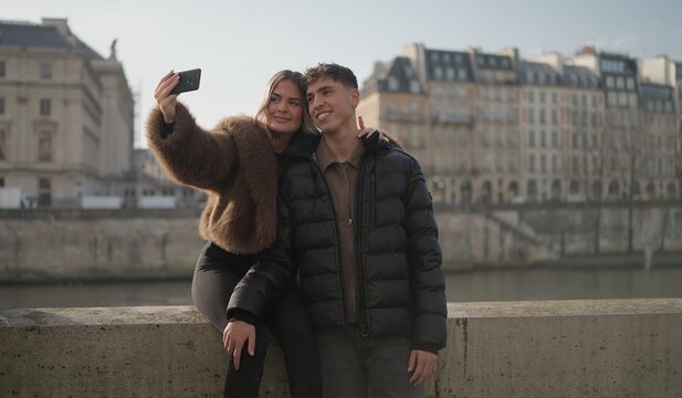 A romantic couple takes a selfie photo with a mobile phone on a bridge over the Seine River in Paris France with city buildings in the background on a sunny day. This image is perfect for travel touri