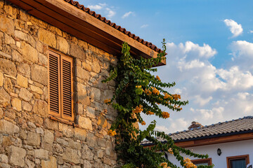 Fototapeta premium Vibrant yellow bougainvillea flowers on a traditional stone Turkish house with wooden shutters, glowing in warm sunset light against a blue sky. Mediterranean, Side, Turkey.