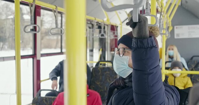 Side view of passengers traveling by public transport. People wearing medical masks, quarantine restrictions, boy in glasses standing, holding bus handle. Concept of covid-19.