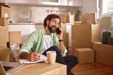 In a modern kitchen filled with moving boxes, a man talks on the phone, jotting down notes while sipping from a coffee cup, balancing tasks during his relocation.