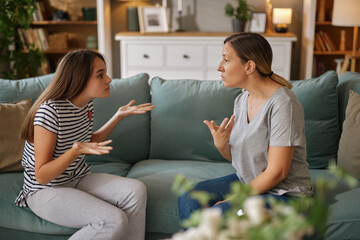 A teenage girl expresses frustration while sitting on a couch, facing her mother who responds calmly. The atmosphere reflects a tense yet loving family moment.