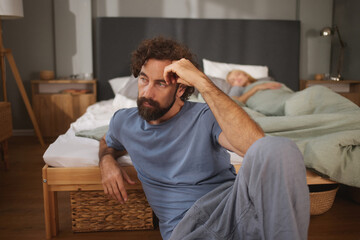 A man sits alone on the floor, thoughtful and worried, while a woman sleeps in bed, highlighting emotional disconnect in their relationship at night.