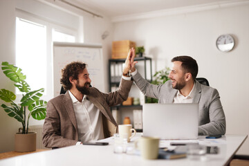 Two team members share a high five in a bright office. They are brainstorming together at a table with a laptop and documents, focused on achieving their goals.