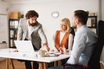 A team of three professionals holds a brainstorming session at a modern office. They discuss strategies while looking at a laptop and taking notes.
