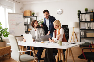 Business team collaborates in an office setting. They share ideas and work together on a project using laptops and notes while seated around a table in a bright space.