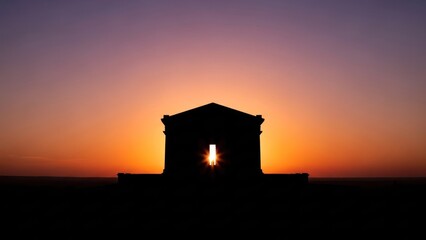 Obraz premium The silhouette of a building against a vibrant sunset sky with a warm orange and purple hue. The structure is a classical architectural style with a pointed roof and arched entrance. 