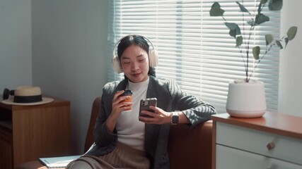 Smiling asian businesswoman relaxing on a couch, enjoying her coffee break while listening to music with headphones and scrolling on her phone