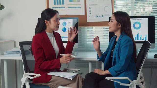 Two professional asian businesswomen discussing financial data and project strategy. Colleagues having a productive conversation in a modern workplace