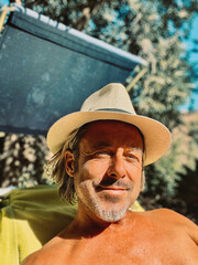 Blond man with straw hat and a stubble beard enjoying sun on a sunbed in garden.