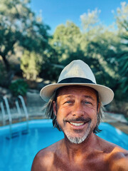 Happy blond man with straw hat and a stubble beard by a pool in a sunny summer garden.