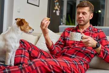 Man wearing pajamas sitting on sofa holding coffee mug and snack while looking at dog