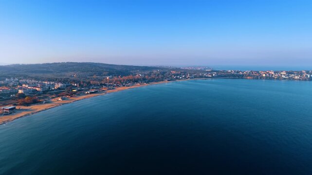 The scene shows a wide view of a beachside town with buildings, green hills, and calm waters during sunset. The sky is clear and the waves are soft along the shore.