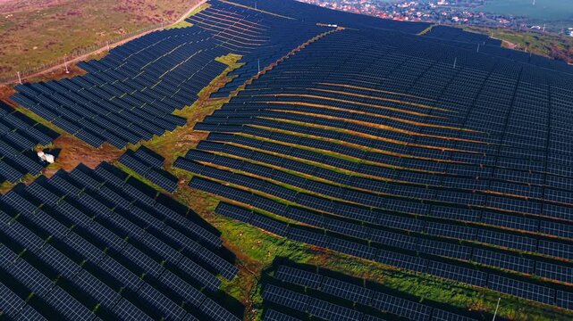 Rows of solar panels cover a large field in a rural area. The panels are aligned neatly. The landscape shows green grass and hills under a clear sky.