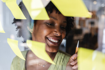 Young African American businesswoman evaluating her creative strategies and new ideas, writing on yellow sticky notes attached to a clear glass wall in a modern office setup. Business concept