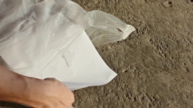 Two hands putting several white paper sheets into a plastic bag on top of a concrete cement floor surface