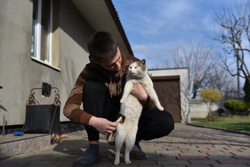 Young man brushing cat outdoors in yard, caring for pet near house on sunny day, calm domestic animal grooming moment © Oleksandr