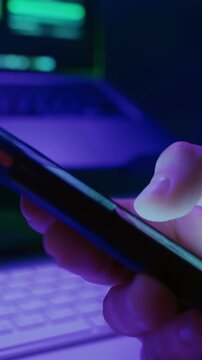 Close-up of persons hand scrolling on smartphone social media feed at night with laptop on background, illuminated by neon light. Digital addiction and modern online habits. Vertical video
