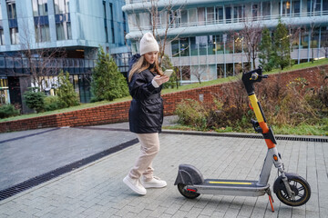 Using smartphone unlocks electric scooter for commute. Activating rental app starts mobility. Standing near parked scooter on wet pavement in rainy weather. Commuter plans route for urban travel.