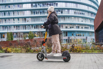 Cruising electric scooter past business district during daily commute. Standing ride posture on wet pavement in rainy weather. Micromobility transport supports fast travel in urban city street.