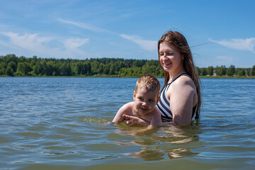Fototapeta premium A smiling young mother teaches her little son how to swim in a lake during the summer. Family outdoor recreation.