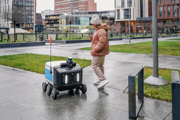 Autopilot delivery robot rolls ahead on wet walkway. Pedestrian follows with smartphone tracking, route guidance. Urban courier logistics enables contactless package dropoff during rainy commute.