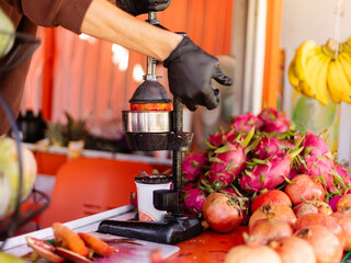 A person is using a hand press to extract juice from pomegranates at a fruit stand. Colorful fruits are displayed around, and the market is busy with shoppers