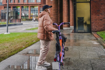 Stopping rented electric bicycle after rainy commute on sidewalk. Holding handlebars during parking, ride finish. Ebike rental sharing supports last mile travel, sustainable mobility, wet route.