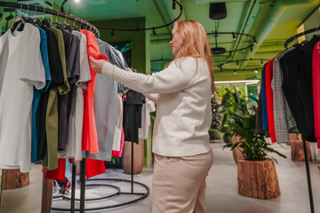 Selecting garment from clothing rack in retail store, shopper compares colors and sizes. Choosing outfit in fashion boutique supports shopping addiction, wardrobe refresh, apparel purchase indoors.
