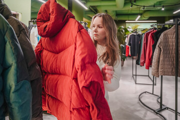Pulling puffer jacket from rack in clothing store, shopper compares outerwear options. Evaluating warm coat for purchase supports seasonal shopping, retail sales, wardrobe refresh indoors today.