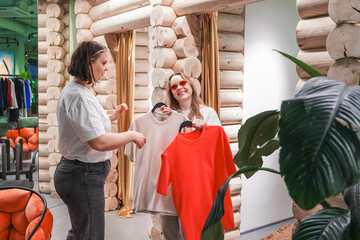 Laughing friends choose clothes in fitting area. Holding garments on hangers, fashion shopping support. Retail boutique lifestyle, purchase planning, wardrobe update, customer experience indoors, joy