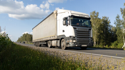 White semi-truck with a cargo trailer moves along an asphalt road through a forest. The truck drives along an empty highway.