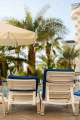 Obraz premium Empty lounge chairs sit by a pool under an umbrella, surrounded by palm trees. The sun shines brightly in the background, creating a relaxing atmosphere at the resort
