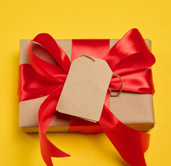 A gift box with a red silk bow and a empty  brown tag on a yellow background, top view