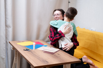 A young woman in a red plaid shirt with braces hugs her son with hearing aids while making a paper garland for Christmas. DIY Christmas home decor.