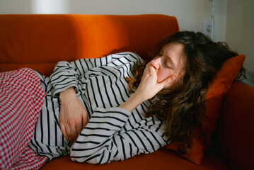 Woman lying on a sofa at home with her hand covering her mouth, feeling nauseous and unwell,...