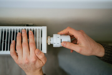 Person's hands adjusting a white thermostatic radiator valve, controlling the room temperature while optimizing energy consumption and reducing high household heating costs