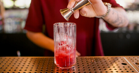 Preparing beverage. Alcohol mixing. Bartender pouring red liquid to glass with ice cubes. Measuring bright cocktail ingredient with jigger.