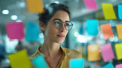 Thoughtful young woman in glasses reflecting on colorful sticky notes in a bright modern office environment with vibrant creativity and brainstorming atmosphere