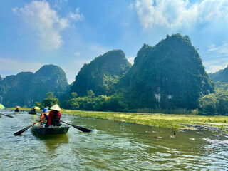 A close-up of the skillful hand-rowing pushing the boat towards majestic mountains. The rower in a red shirt stands out against the green water © Bachhuyhoang
