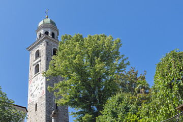 Obraz premium Stone Clock Tower With Green Dome And Cross Under Clear Blue Sky Surrounded By Trees, Lugano Cathedral, Cattedrale di San Lorenzo, Lugano, Switzerland