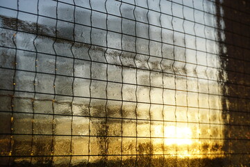 Abstraktes Motiv mit Glasscheibe und Gitterk&auml;stchenmuster vor goldgelb-grau-blauem Himmel mit wei&szlig;em Licht bei Frost, Eis und K&auml;lte bei Sonnenaufgang am Morgen im Winter