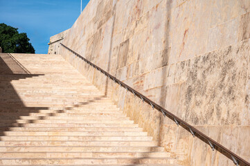 Fototapeta premium Historic ochre-colored stone stairs at the Parliament of Valletta, showcasing geometric patterns and shadows Valletta, Malta, 6 DEC 2025.