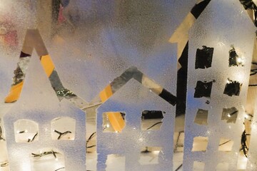 Weihnachtsdekoration an Fensterscheibe mit wei&szlig;er aufgeklebter H&auml;userreihe in Schneelandschaft mit Licht am Abend im Winter