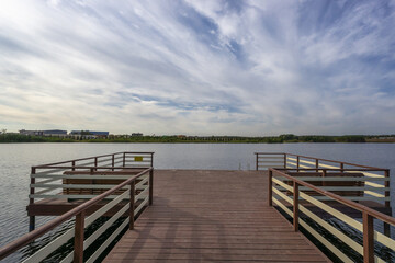 Fototapeta premium A wooden pier reaches out over still water. Trees and buildings line the far shore under a cloudy sky. The scene shows a quiet outdoor space for walking or fishing.